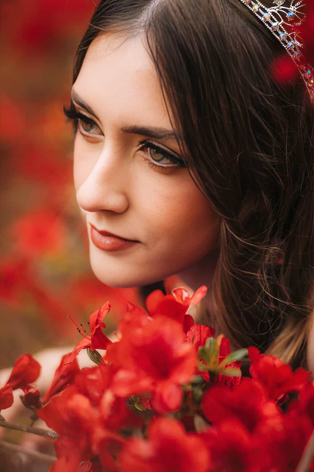 Close-up of a quinceañera in a crystal tiara gazing past a cluster of red azaleas