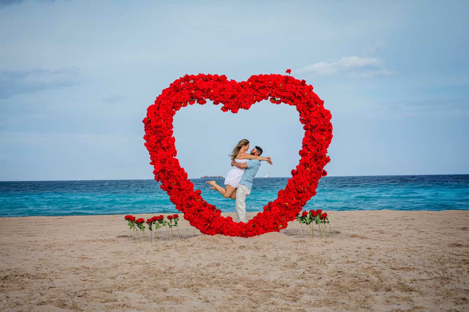He lifts her in celebration inside a giant heart of red roses on the beach after a surprise proposal