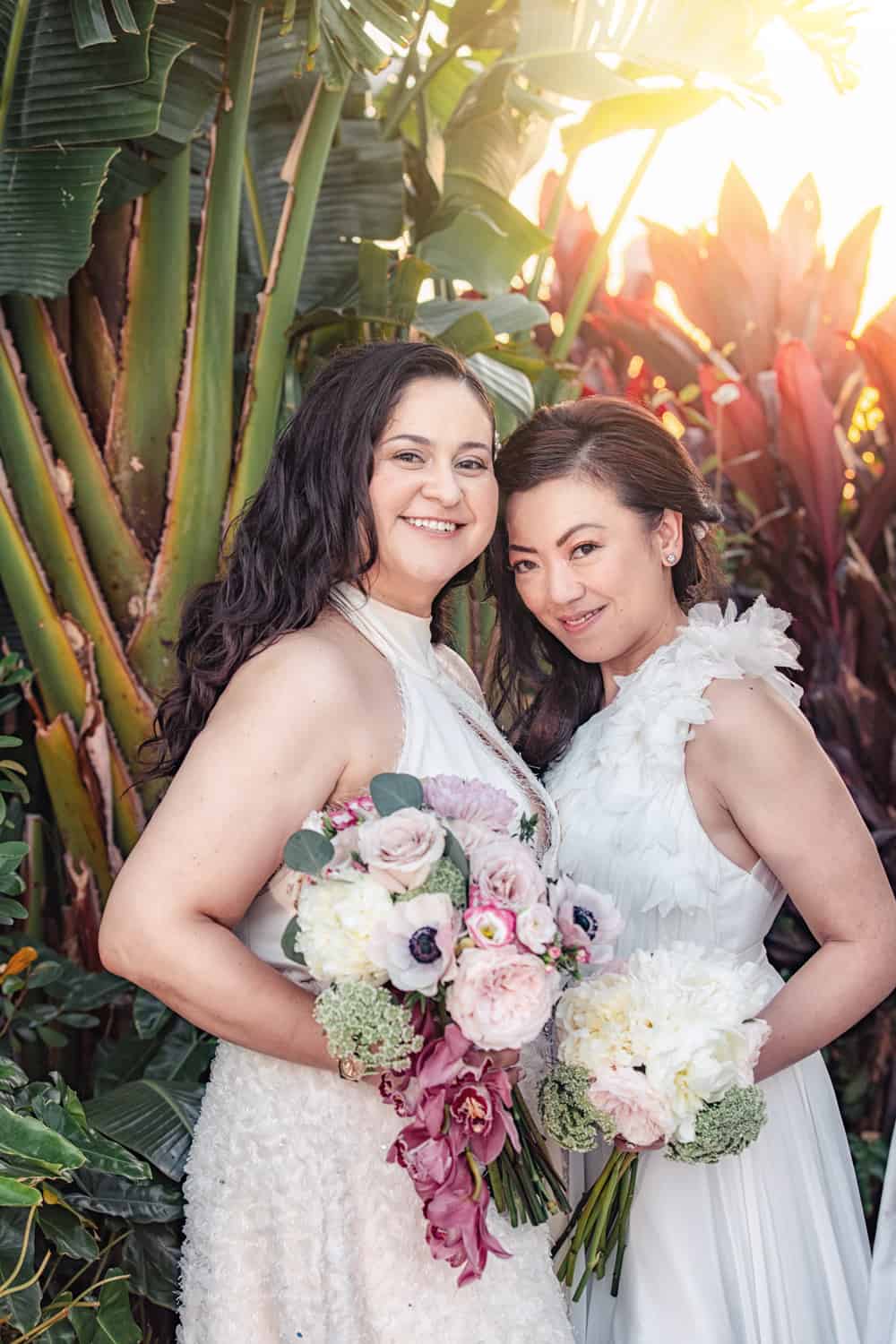 Two brides with blush and white bouquets smiling in a tropical garden at golden hour, The Ray Hotel, Delray Beach