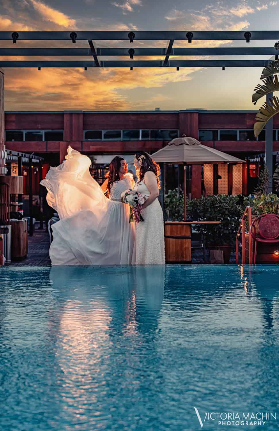 Two brides face to face by a rooftop pool at sunset, a veil lifting in the wind, The Ray Hotel, Delray Beach