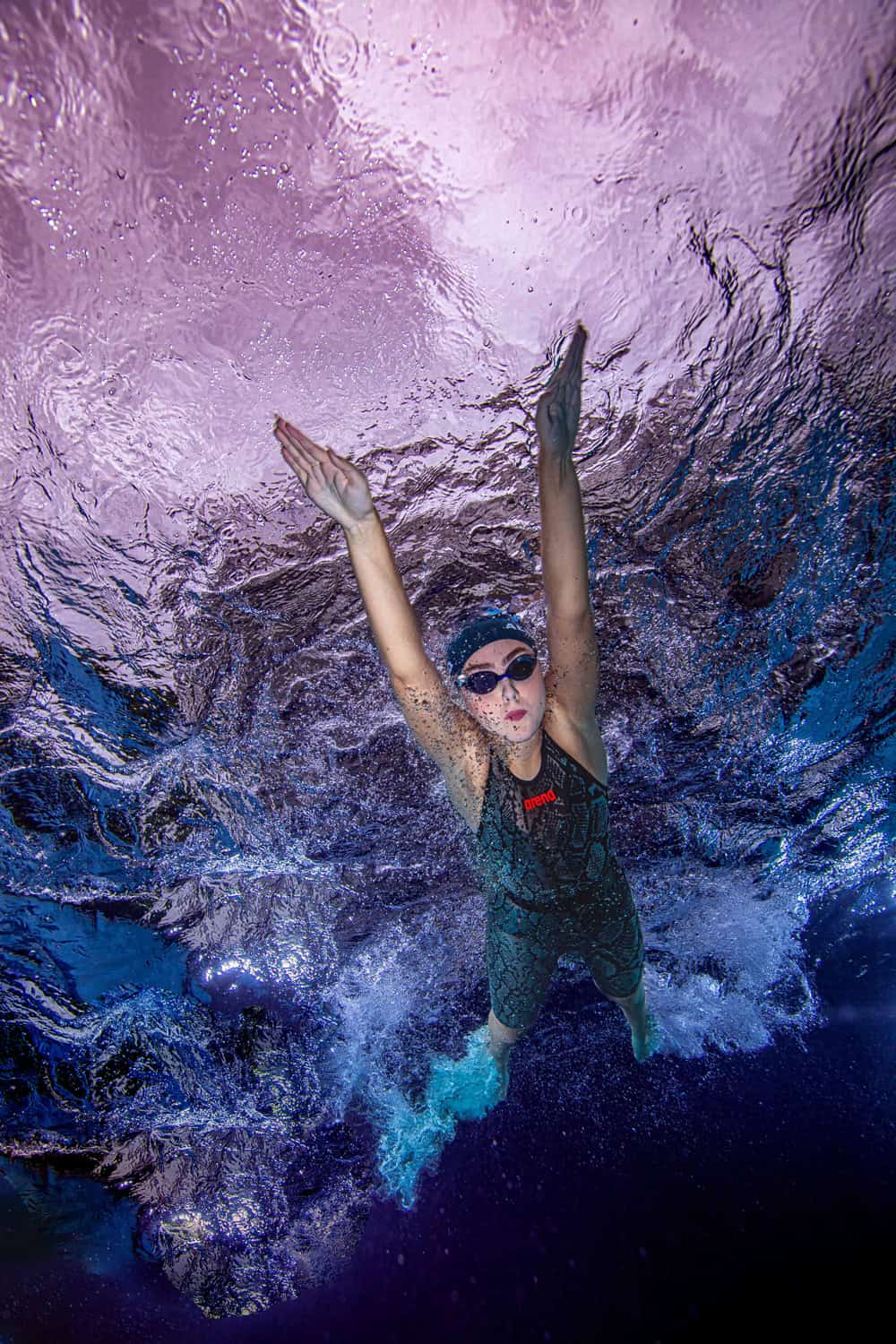 Competitive swimmer diving down through bubbles beneath a violet surface
