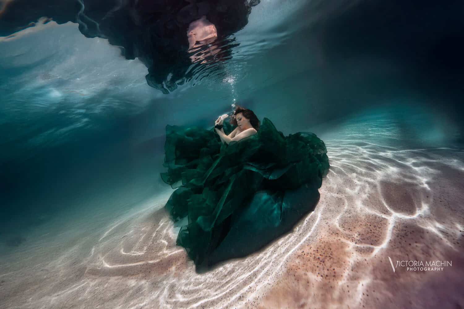 Woman in an emerald gown resting on the pool floor beneath dark water and rippling light