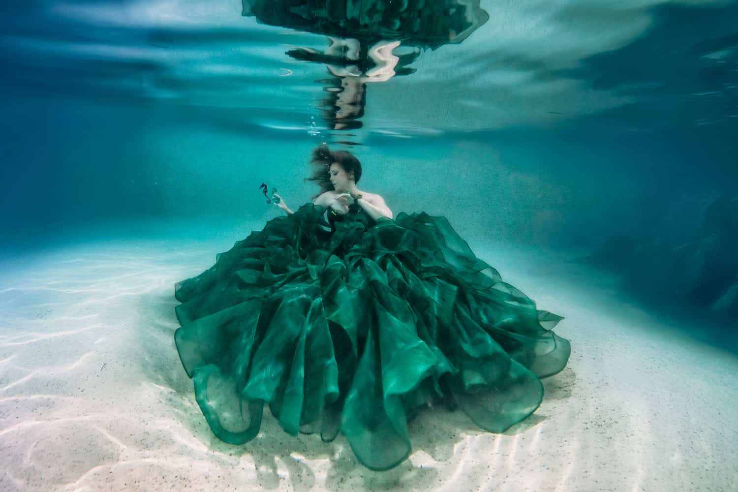 Underwater fine-art portrait: a woman in an emerald gown resting on white sand beneath the surface