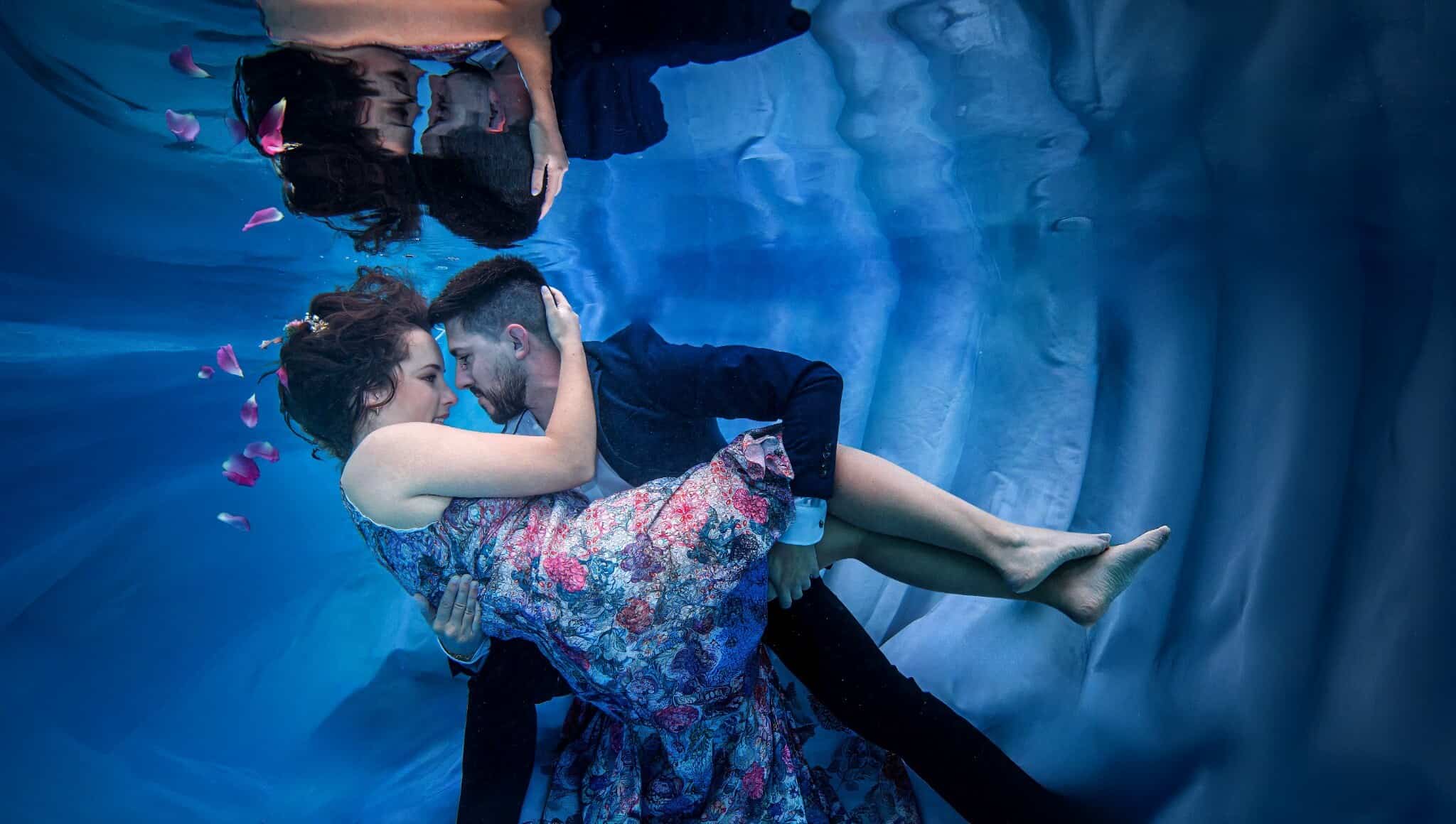 Groom lifting his bride-to-be underwater as rose petals drift past