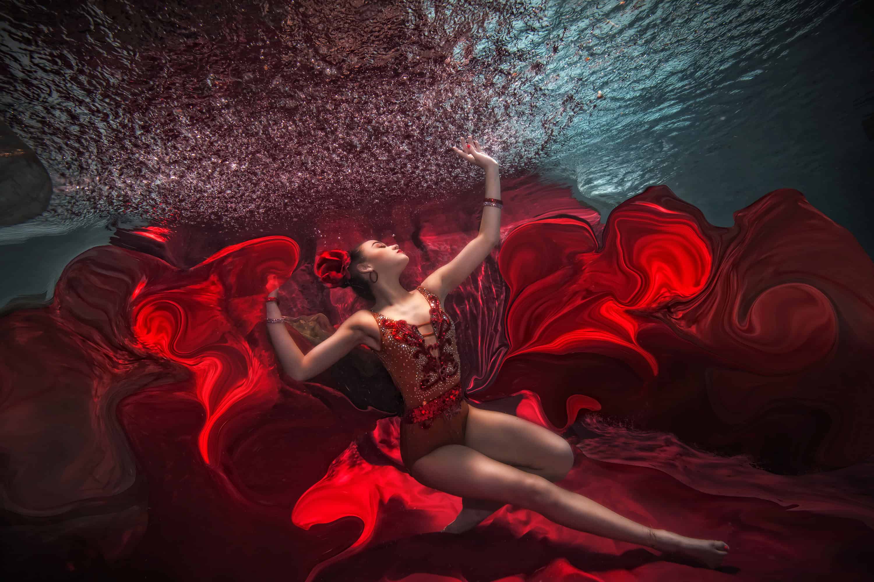 Dancer in a red-embellished leotard arched among swirling red fabric underwater