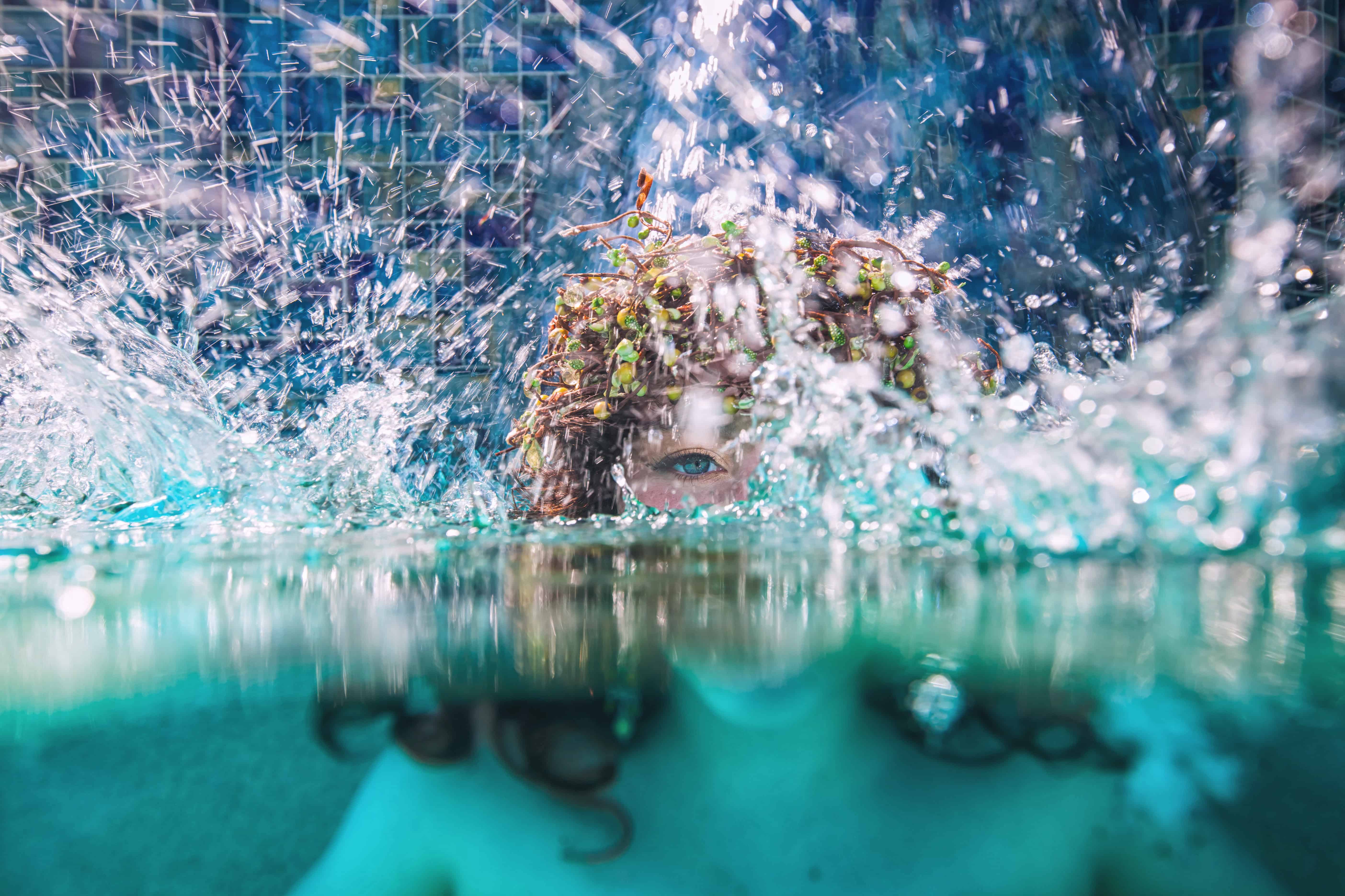 Half-above, half-below portrait of a girl in a floral crown breaking the surface in a burst of spray