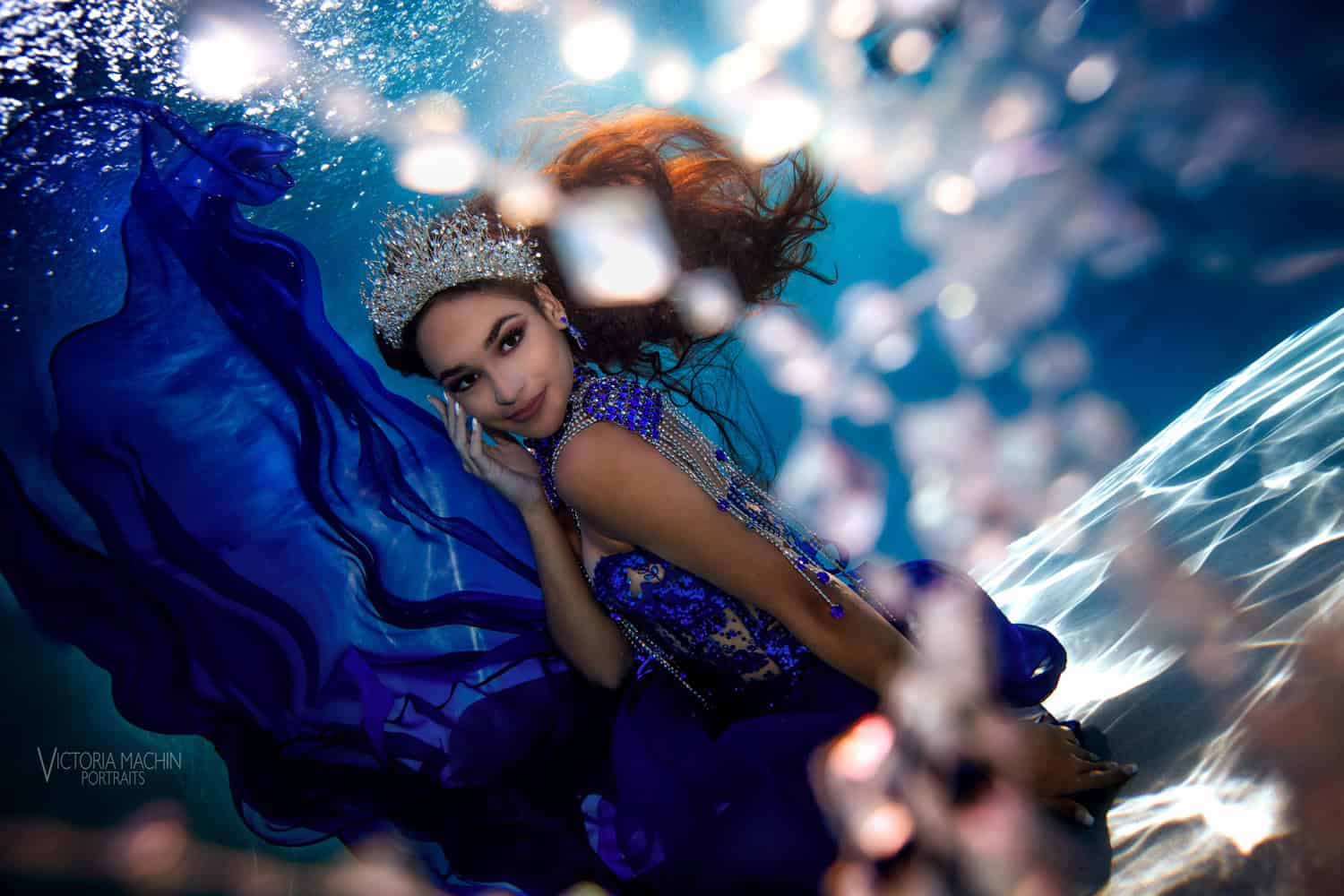 Quinceañera in a crystal crown and royal blue gown smiling through bubbles and light