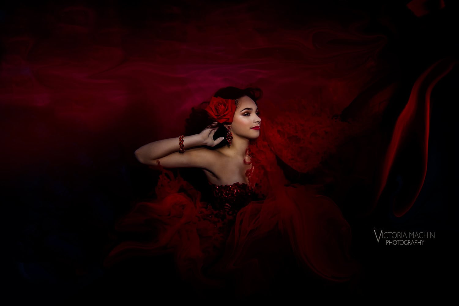 Quinceañera with a red rose in her hair in a red gown against dark water
