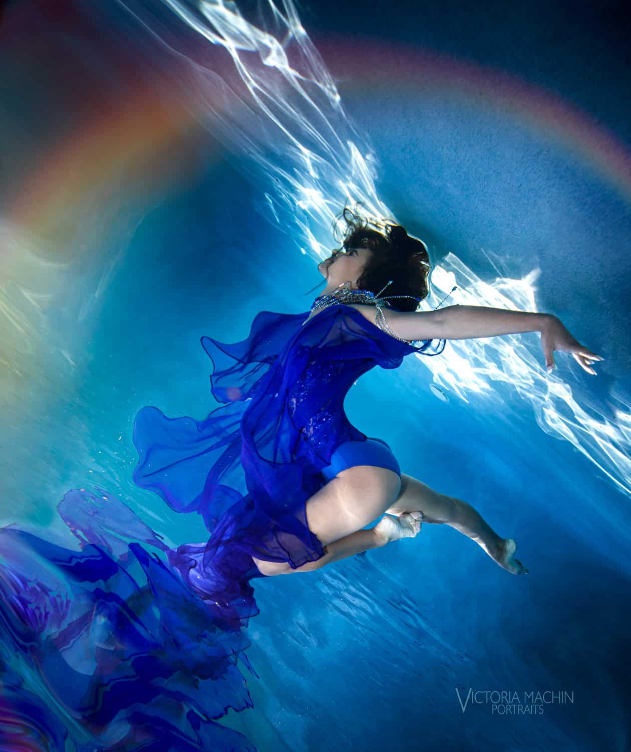 Quinceañera in a royal blue gown soaring through sunlit water with a rainbow of light above