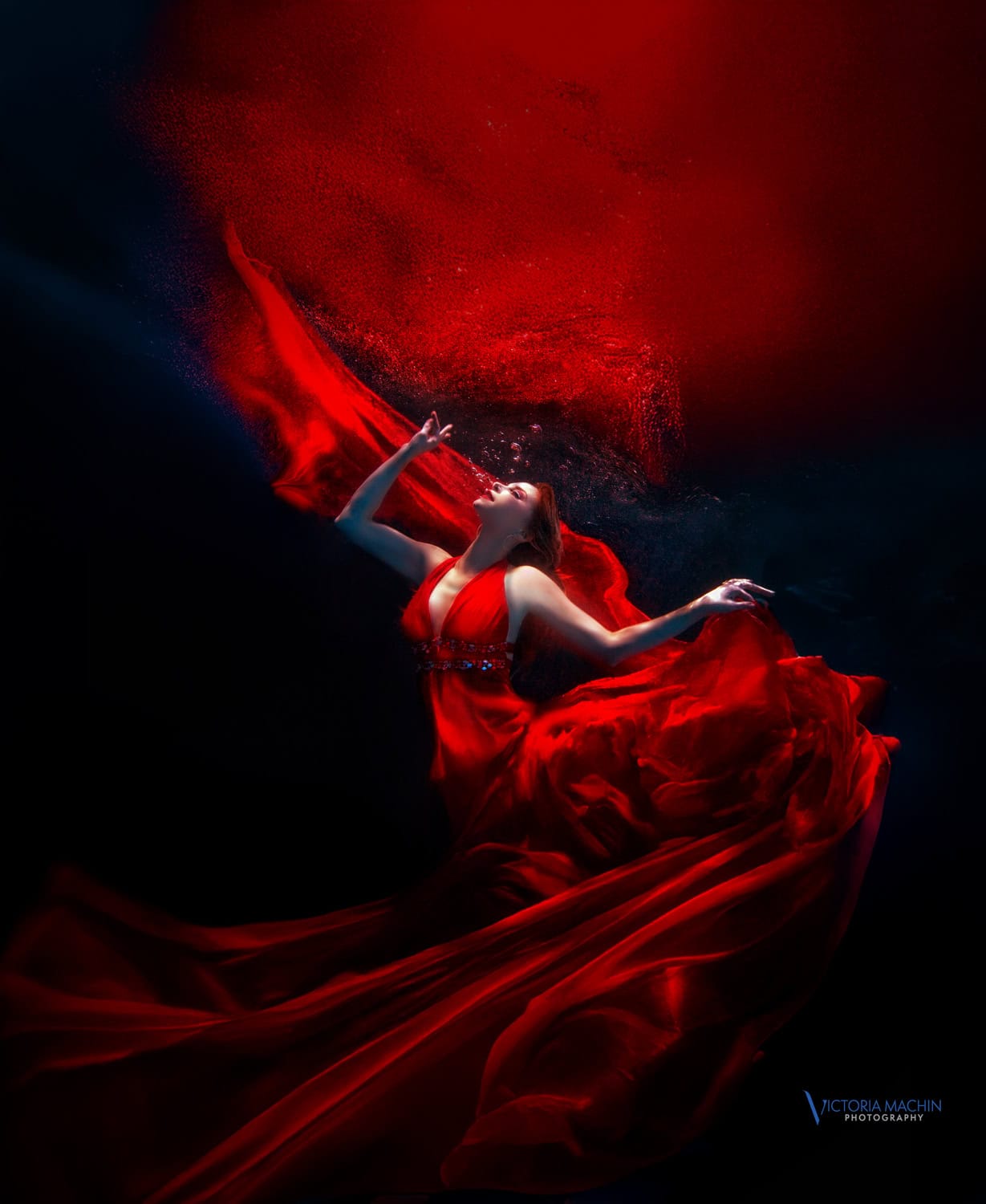 Woman in a flowing red gown arching through black water, fabric trailing above her