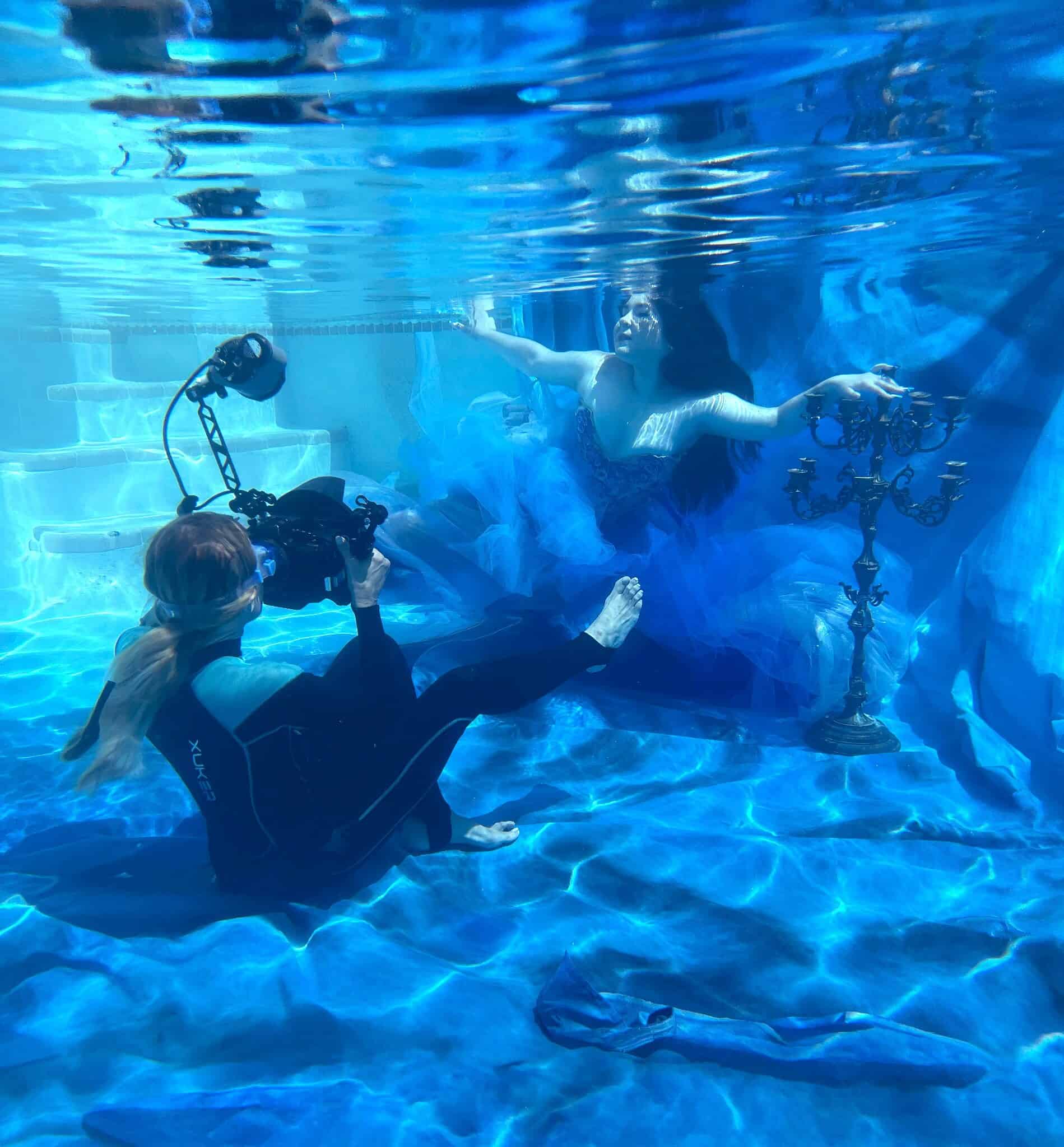 Victoria Machin in a wetsuit photographing a quinceañera in a blue gown underwater beside a candelabra