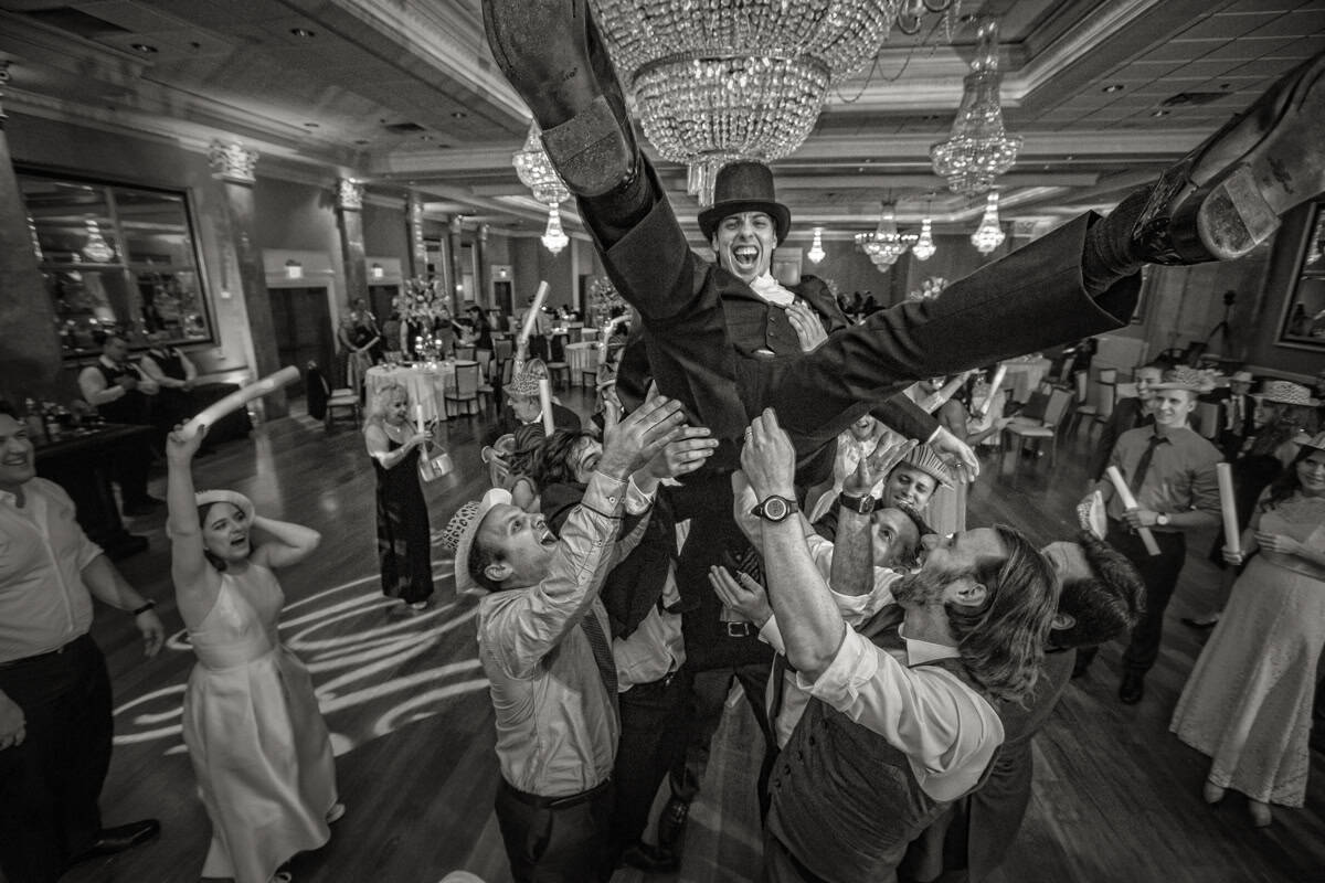Groom in a top hat lifted high by guests on a crowded reception dance floor, in black and white